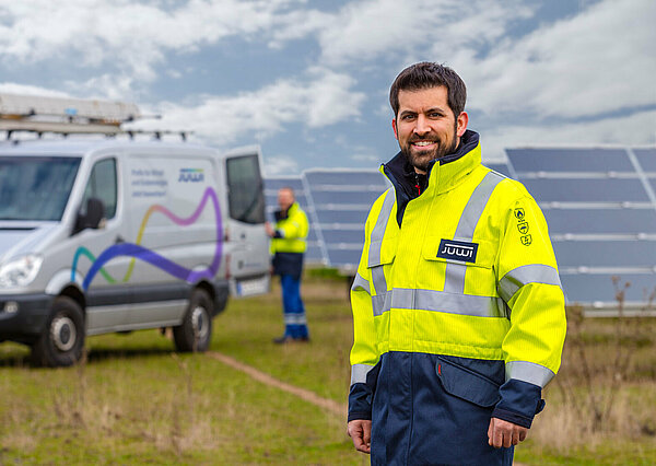 A JUWI colleague in a neon-colored jacket is smiling at the camera. In the background, another colleague is working on a solar module.